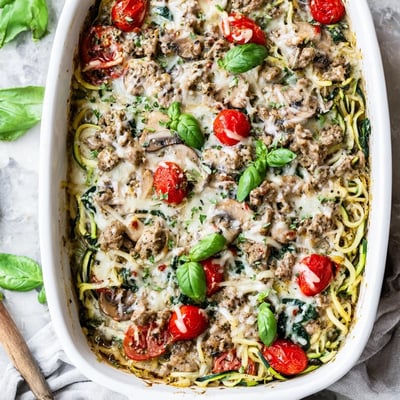 Creamy low carb pesto turkey and mushroom zoodle bake topped with browned cheese in a white casserole dish